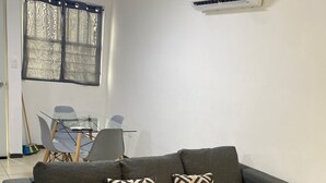Ένα σαλόνι με έναν γκρι γωνιακό καναπέ, δύο διακοσμητικά μαξιλάρια με σχέδια, ένα τραπεζάκι σαλονιού από γυαλί και ένα τραπέζι τραπεζαρίας με καρέκλες. Μια μονάδα κλιματισμού είναι τοποθετημένη στον τοίχο πάνω από τον καναπέ.