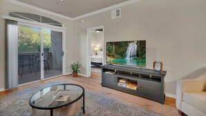 A living room with a TV, a glass coffee table, a sofa, a sliding glass door leading to a balcony, and a ceiling fan.