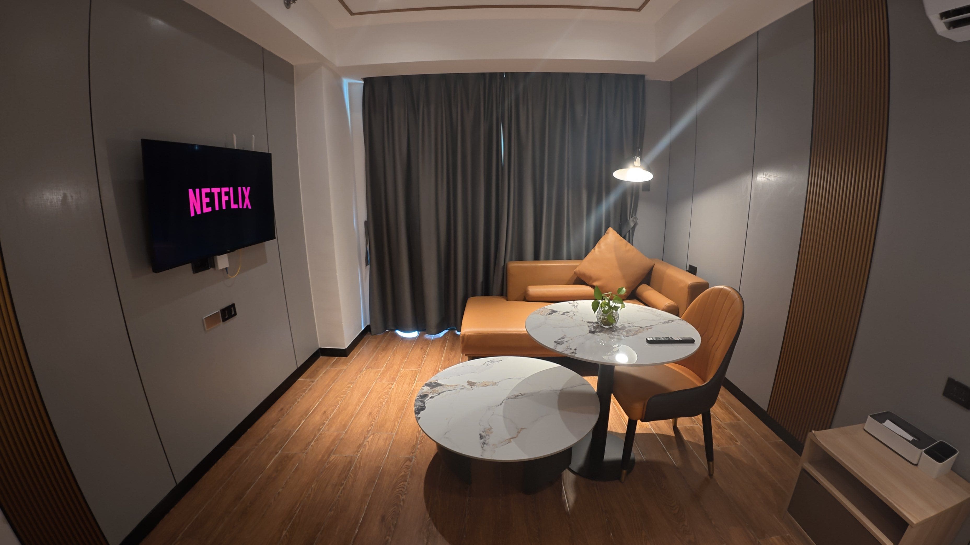A modern hotel room with a flat-screen TV displaying 'NETFLIX,' a leather sofa, a round coffee table, and a small side table with a remote control.
