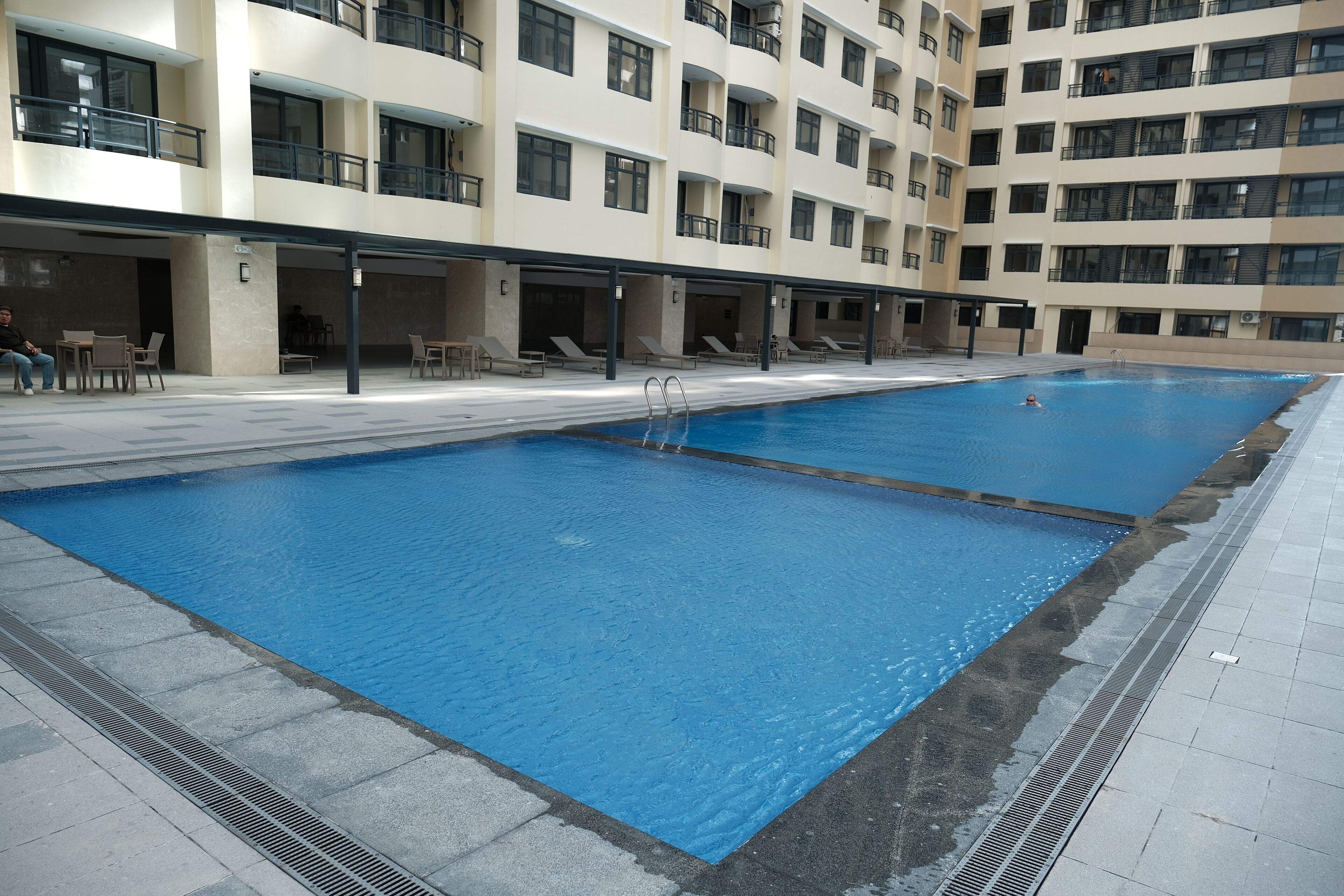 A large indoor swimming pool with clear blue water, surrounded by a tiled deck and a multi-story building with balconies.