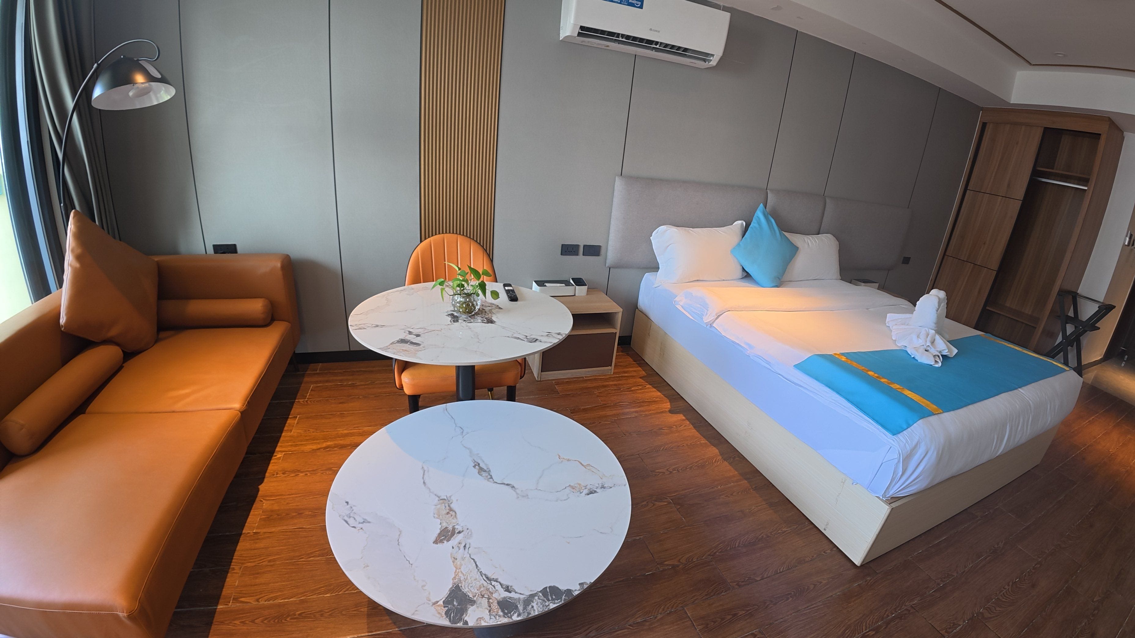 A modern hotel room with a sofa, a round table, a bed with a blue bedspread, and an air conditioning unit.