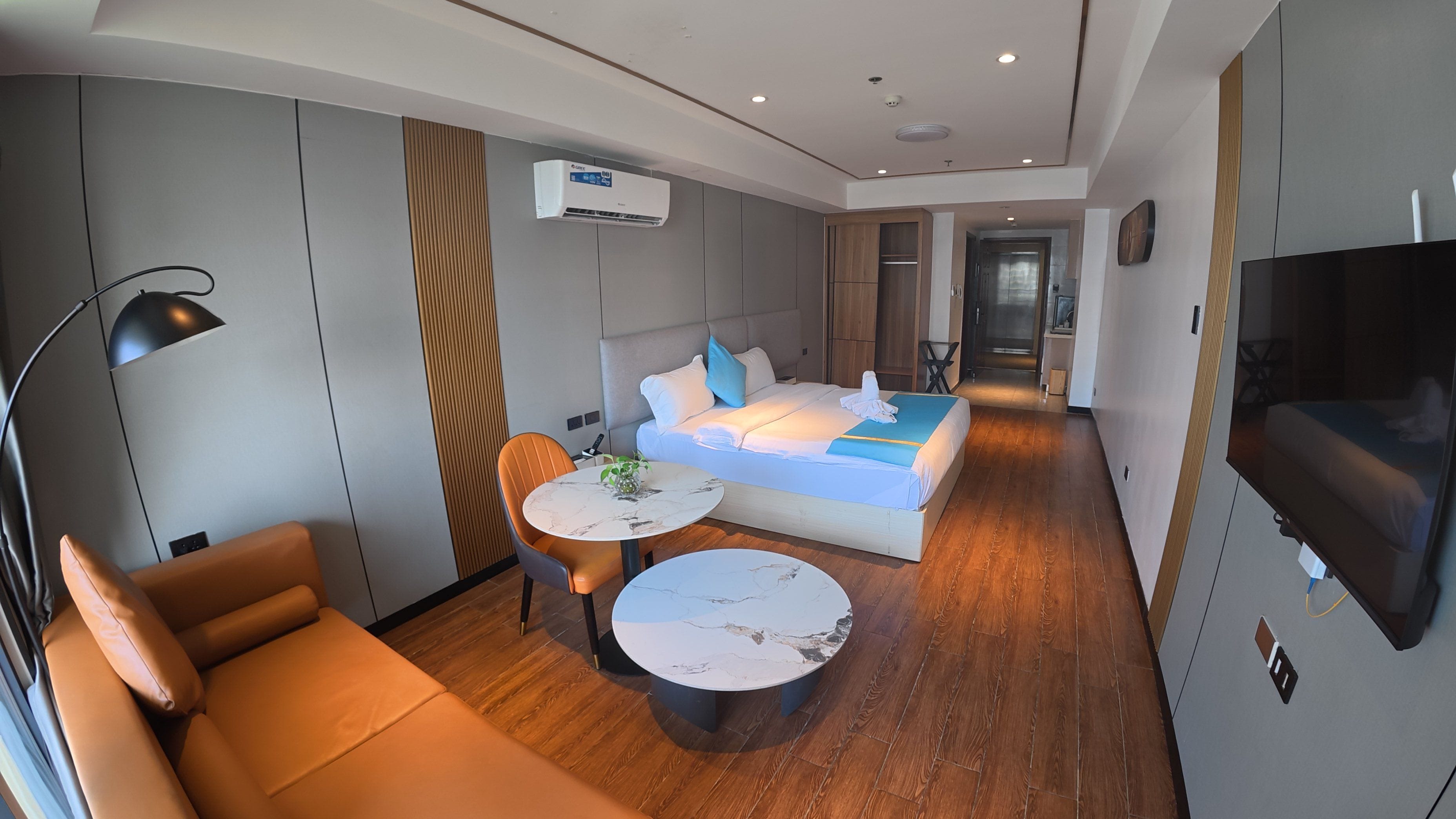 A modern hotel room with a sofa, a bed, a TV, a desk, and a wall-mounted air conditioner.