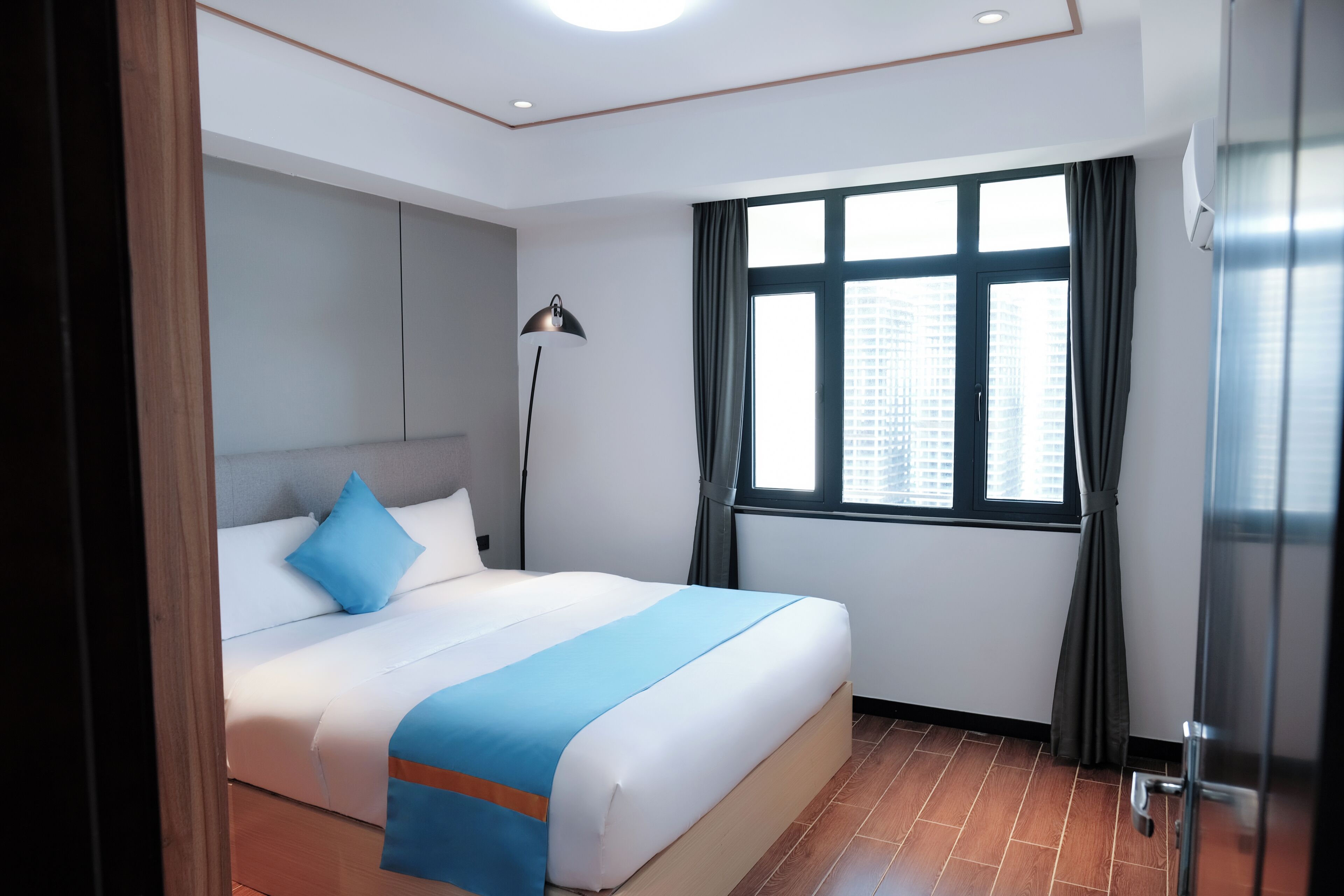 Habitación de hotel con una cama grande, una lámpara de pie, un ventanal con cortinas y vistas a edificios.