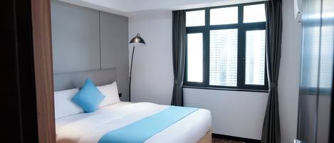 Habitación de hotel con una cama grande, una lámpara de pie, un ventanal con cortinas y vistas a edificios.