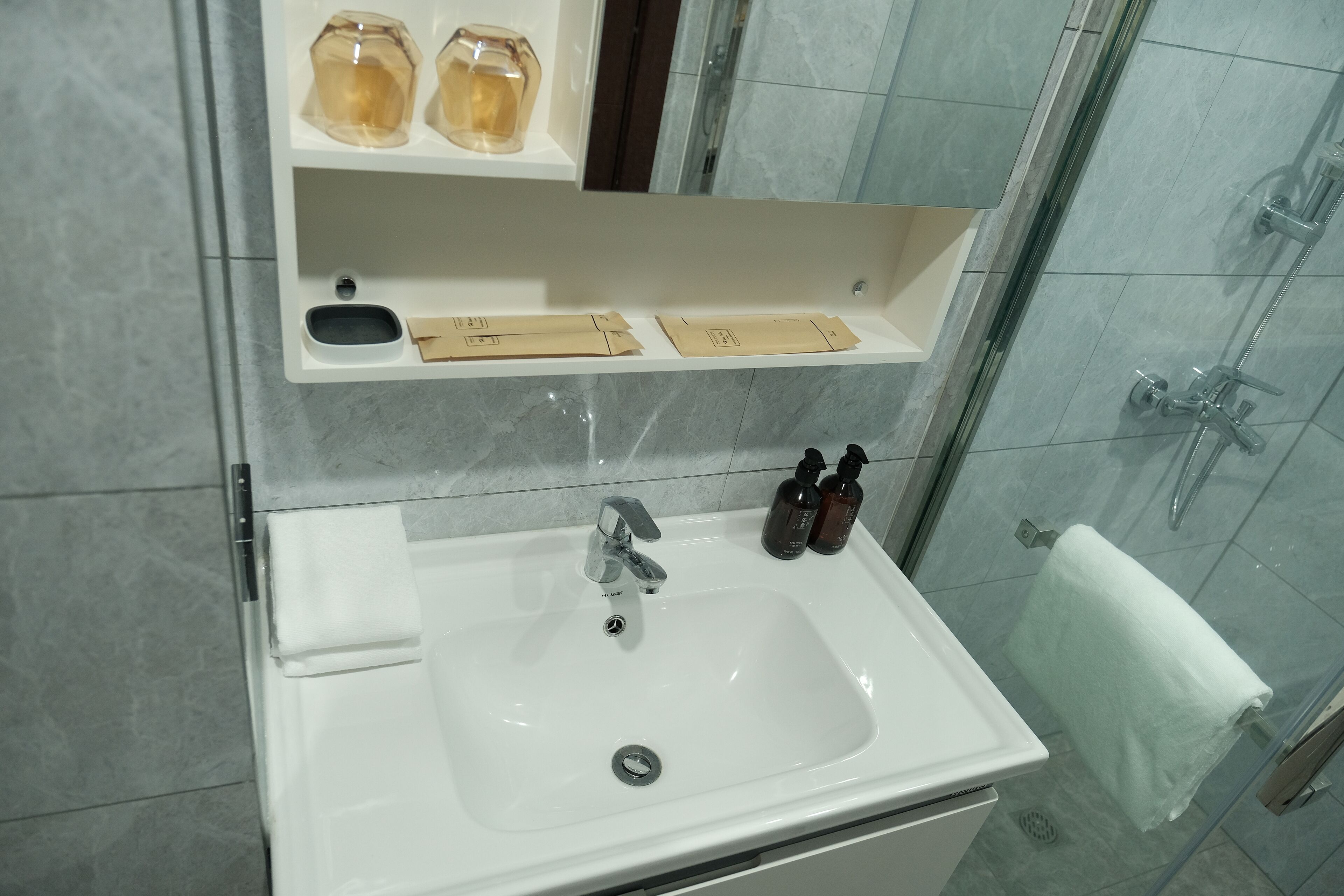 Ein Badezimmer mit einem weißen Waschbecken, einem Spiegel darüber und einer Dusche rechts.