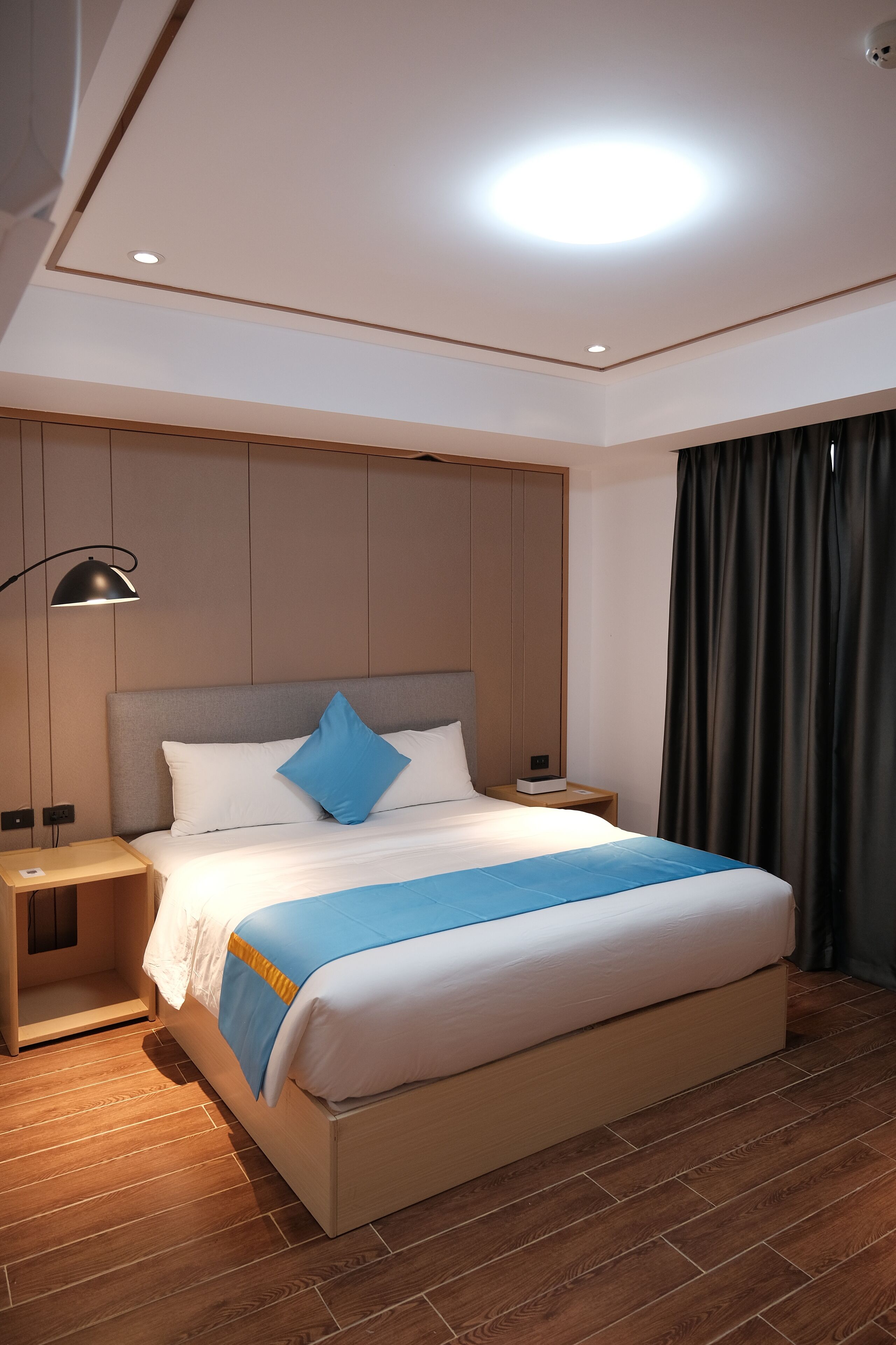 A modern hotel room with a large bed, wooden flooring, a nightstand, a lamp, and a ceiling light.
