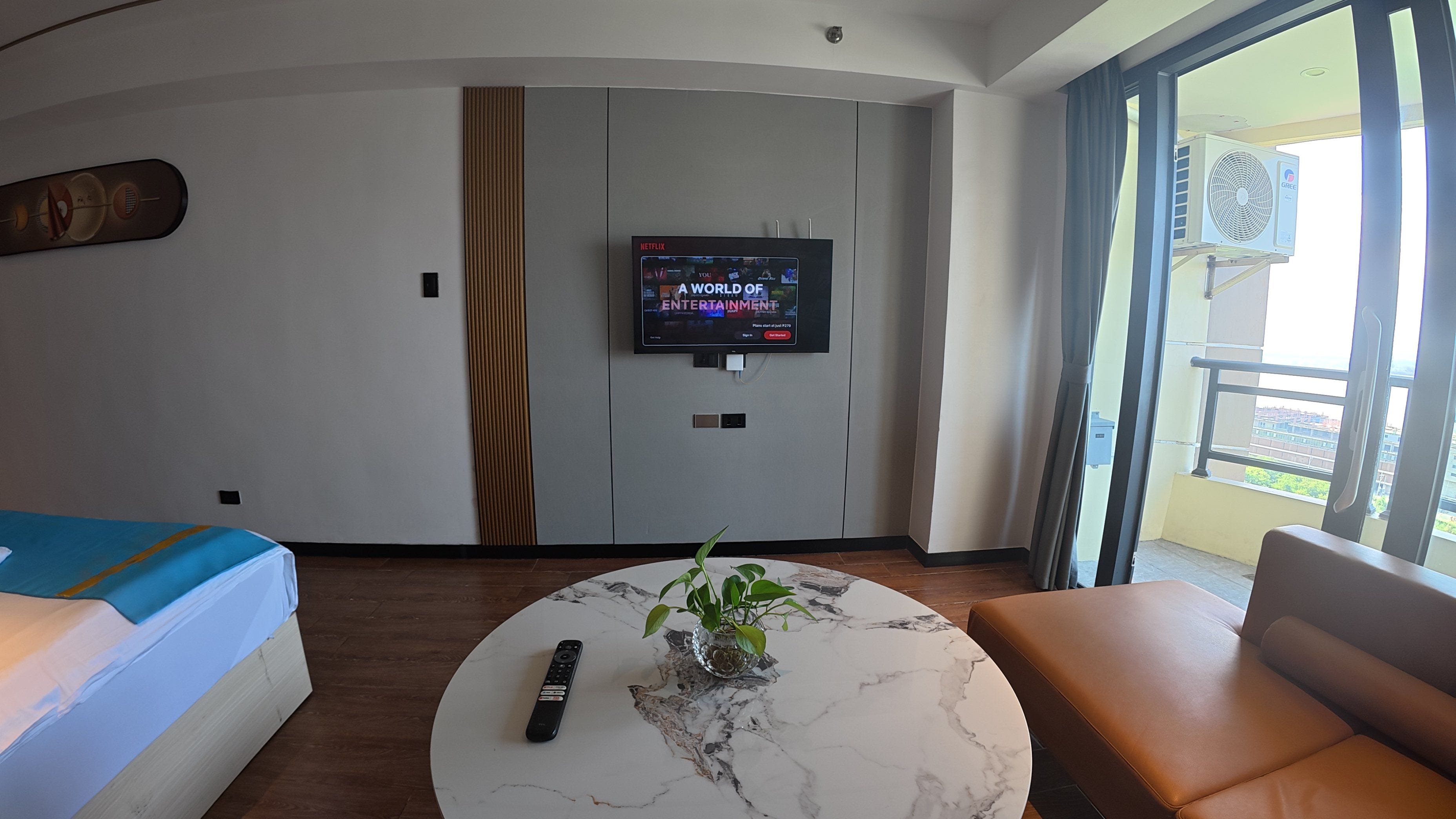 A hotel room with a bed, a sofa, a coffee table with a plant, a TV on the wall, and a balcony with an air conditioning unit.