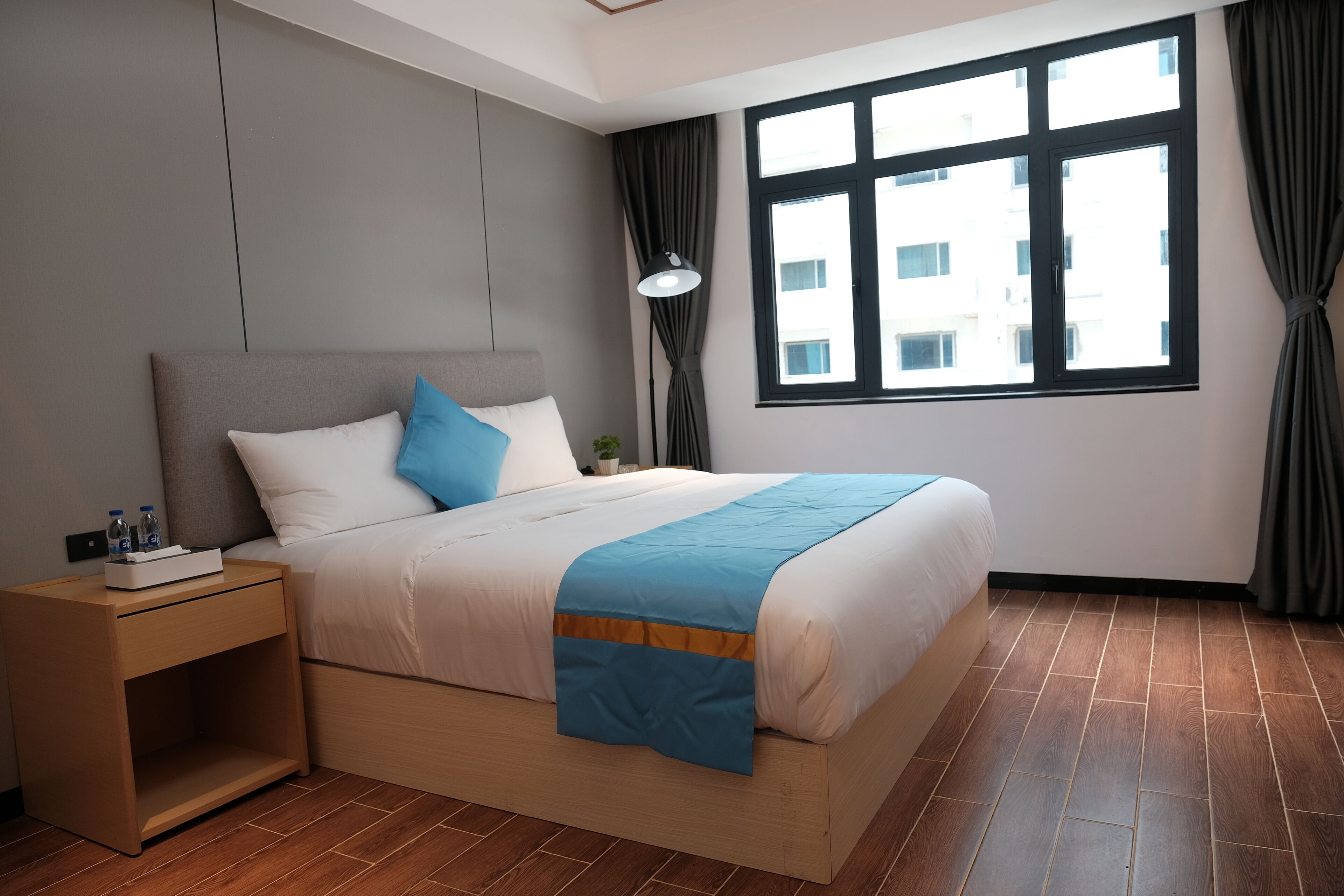 Una habitación de hotel moderna con una cama de madera, una mesita de noche, una ventana con cortinas y vistas a edificios.