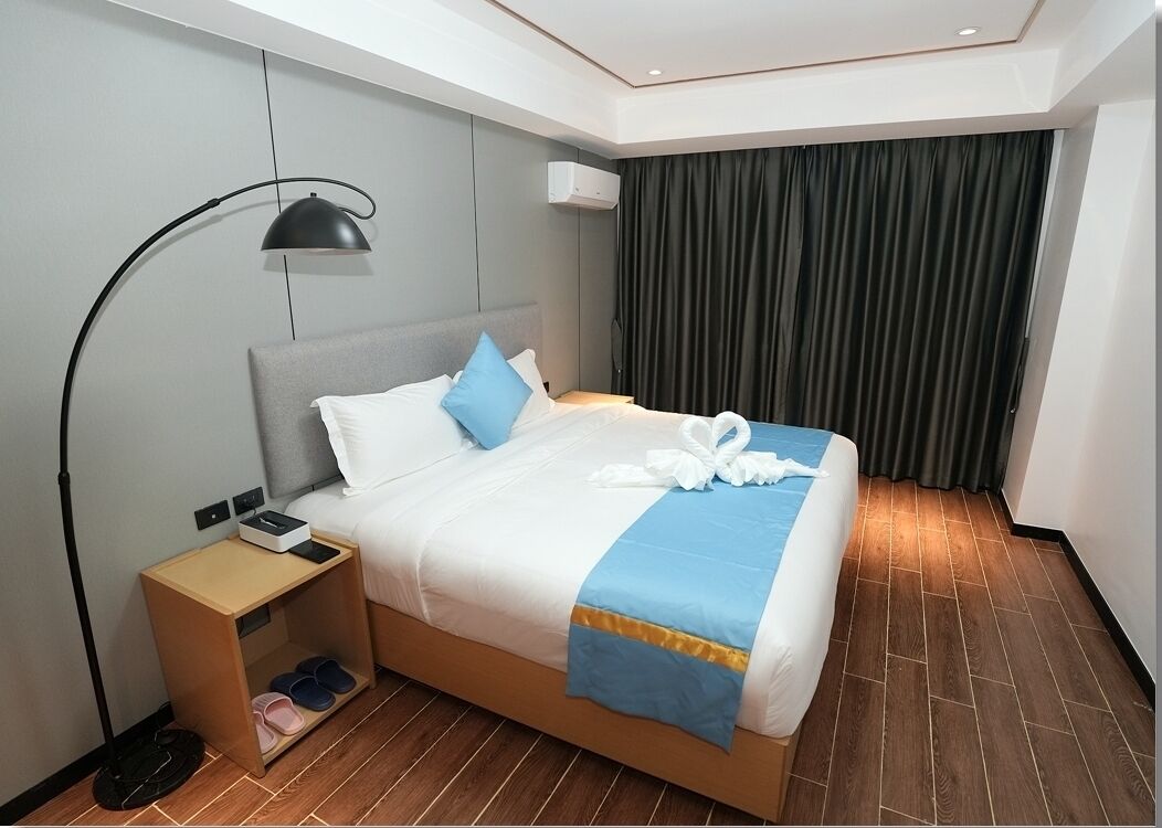 Une chambre d’hôtel avec un lit, une table de chevet, une lampe, un porte-chaussures et un rideau.