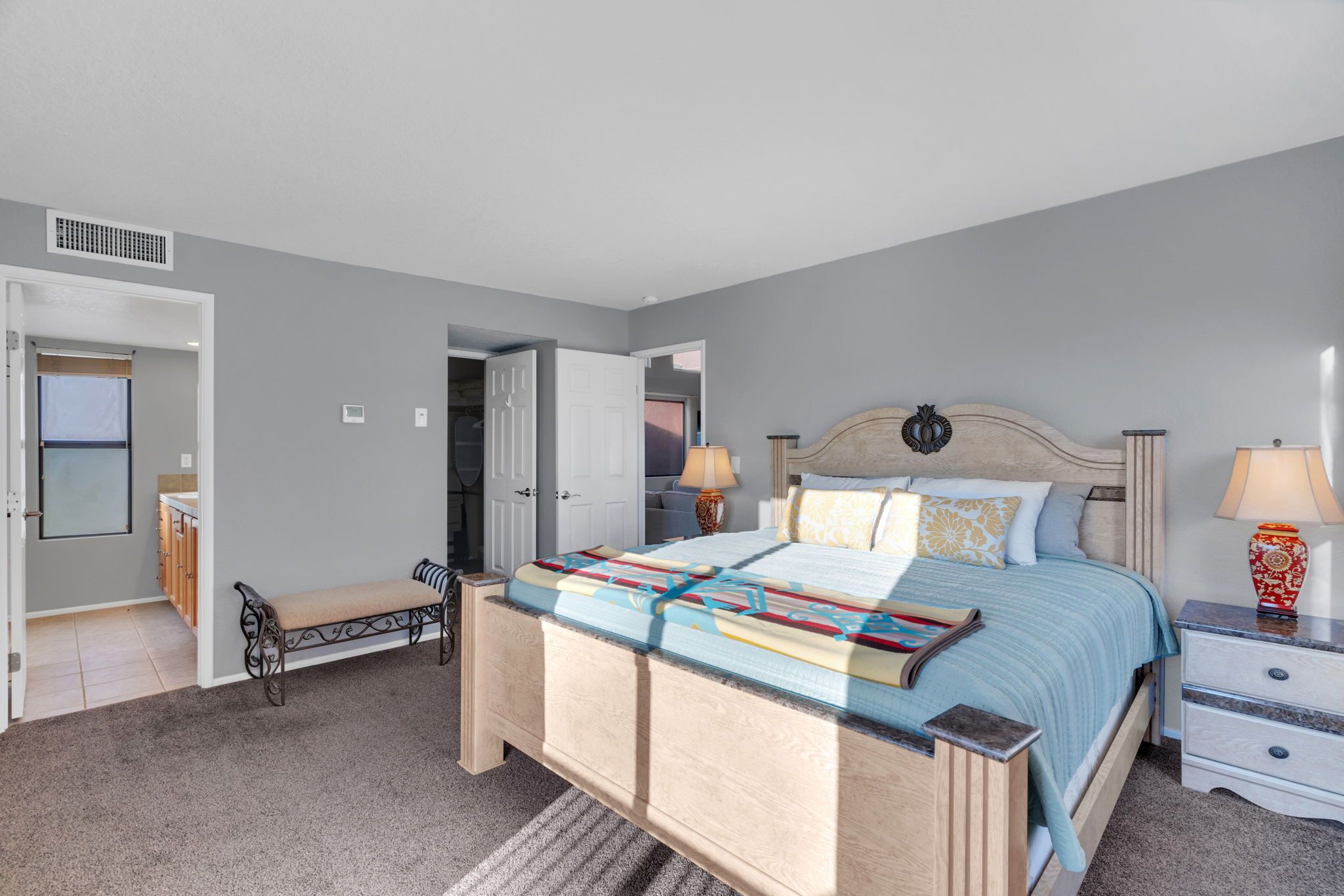 Quarto com cama, mesas de cabeceira, um banco e uma porta que dá para outro cômodo.