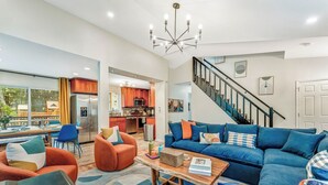 Un vaste espace de vie comprenant un canapé d’angle bleu, des fauteuils orange et une table basse en bois. Un escalier avec une rampe noire mène à l’étage supérieur.