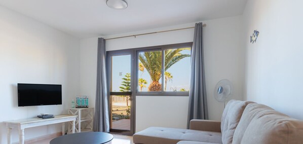 Ein modernes Wohnzimmer mit Flachbildfernseher, einer Couch, einem Couchtisch und einer Schiebetür zu einem Balkon mit Blick auf eine Palme.