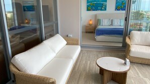 A modern room with a white sofa, a round wooden table, and a bed with a blue and white striped bedspread. There are two paintings on the wall depicting ocean scenes.