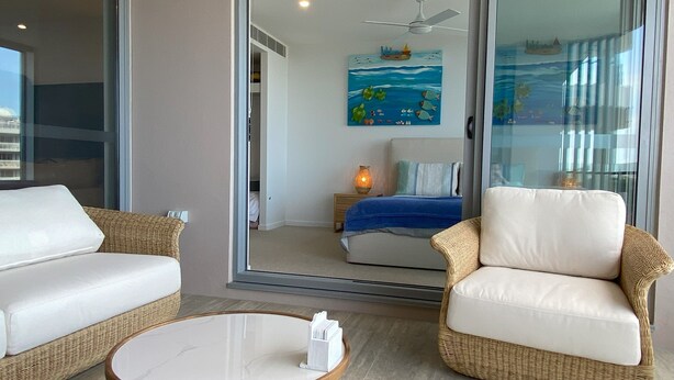 A modern living room with a glass door leading to a bedroom. The room features a round wooden coffee table, two wicker armchairs, a white sofa, and a bed with a blue blanket. A ceiling fan and a wall painting of a seascape are visible.
