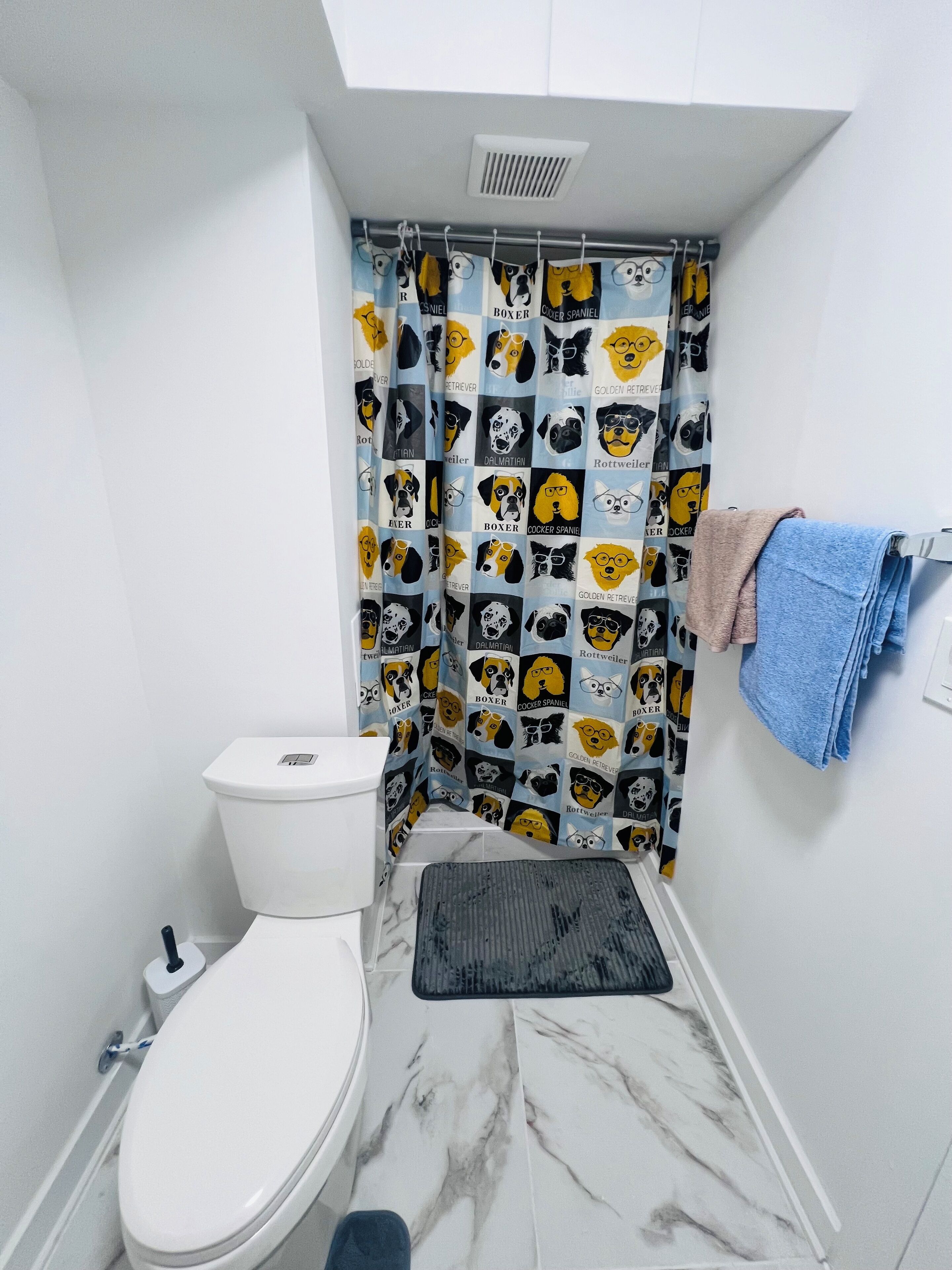 A bathroom with a toilet, a shower curtain with dog prints, a towel rack with towels, and a marble floor.