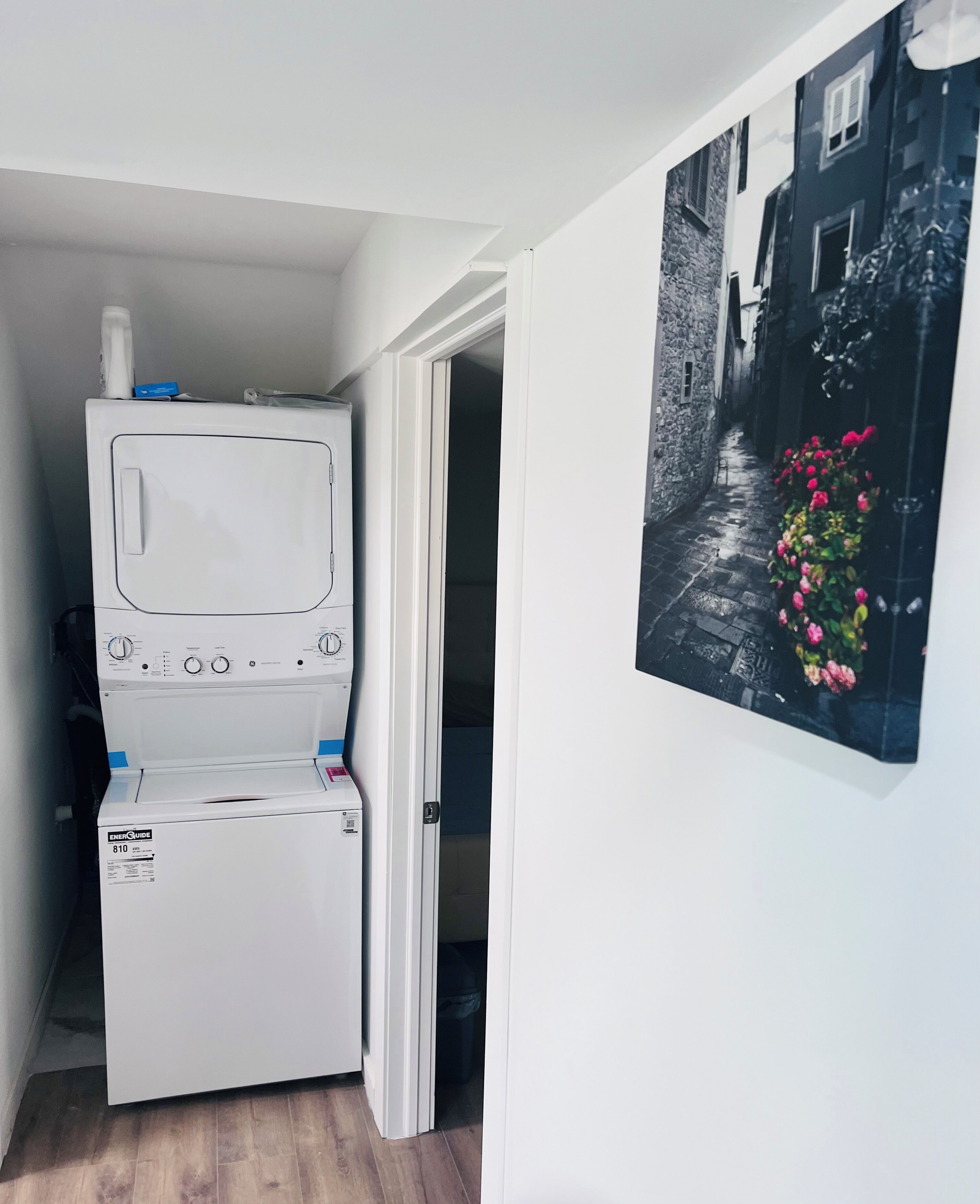 A compact laundry room with a front-loading washer and dryer, a door leading to a narrow hallway, and a wall-mounted picture of a street scene with flowers.