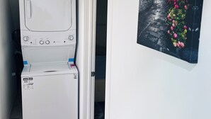 A compact laundry room with a front-loading washer and dryer, a door leading to a narrow hallway, and a wall-mounted picture of a street scene with flowers.