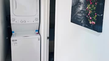 A compact laundry room with a front-loading washer and dryer, a door leading to a narrow hallway, and a wall-mounted picture of a street scene with flowers.