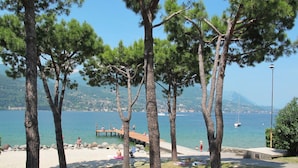 A beach with people sunbathing and relaxing under trees, a wooden pier extending into the water, and a distant view of a mountain. - Vacation home Eden by Interhome (San Felice del Benaco)