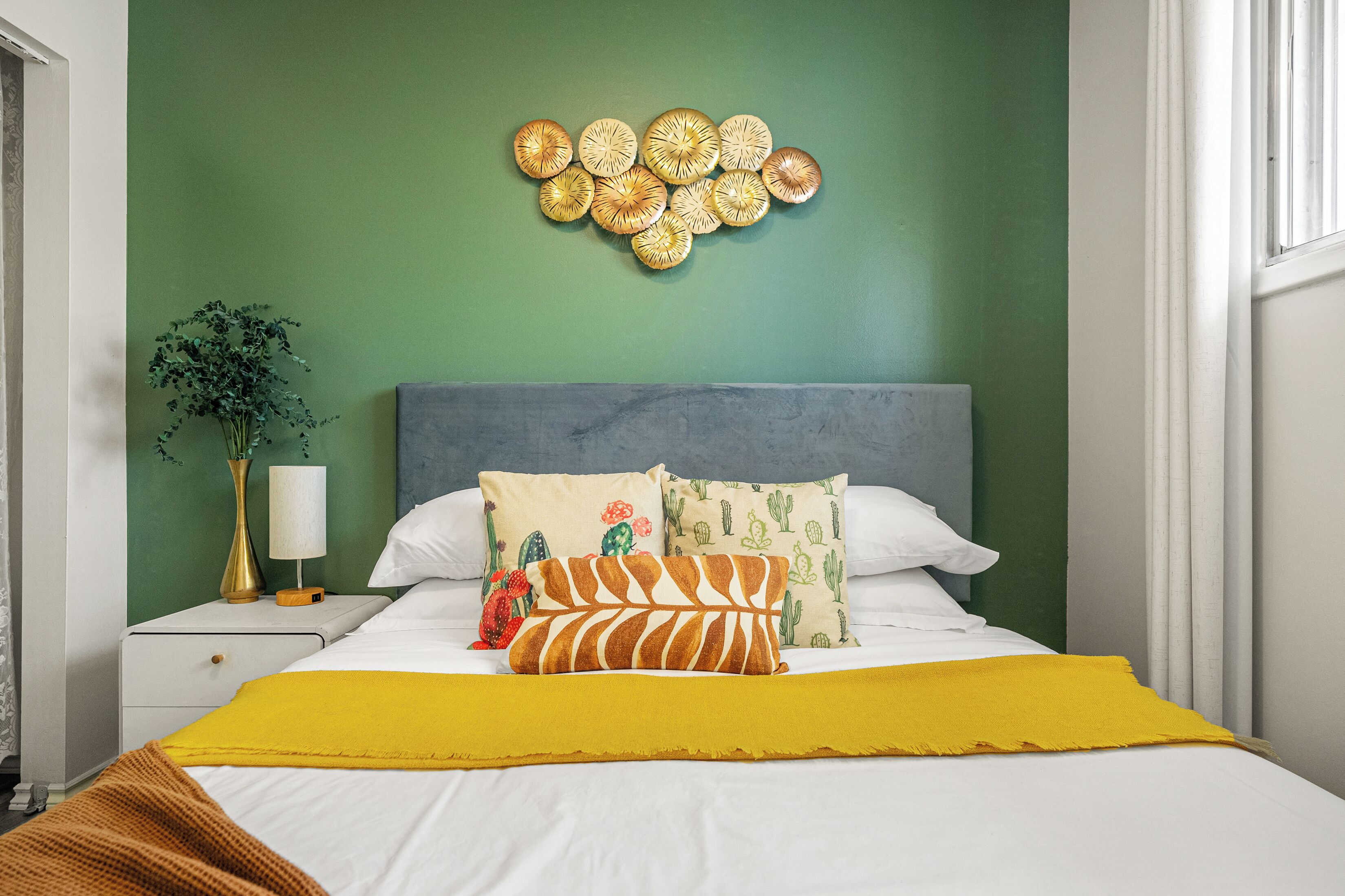 Un lit bien fait, agrémenté d’une couverture jaune, d’un coussin à motifs de cactus et d’une œuvre d’art murale au-dessus.