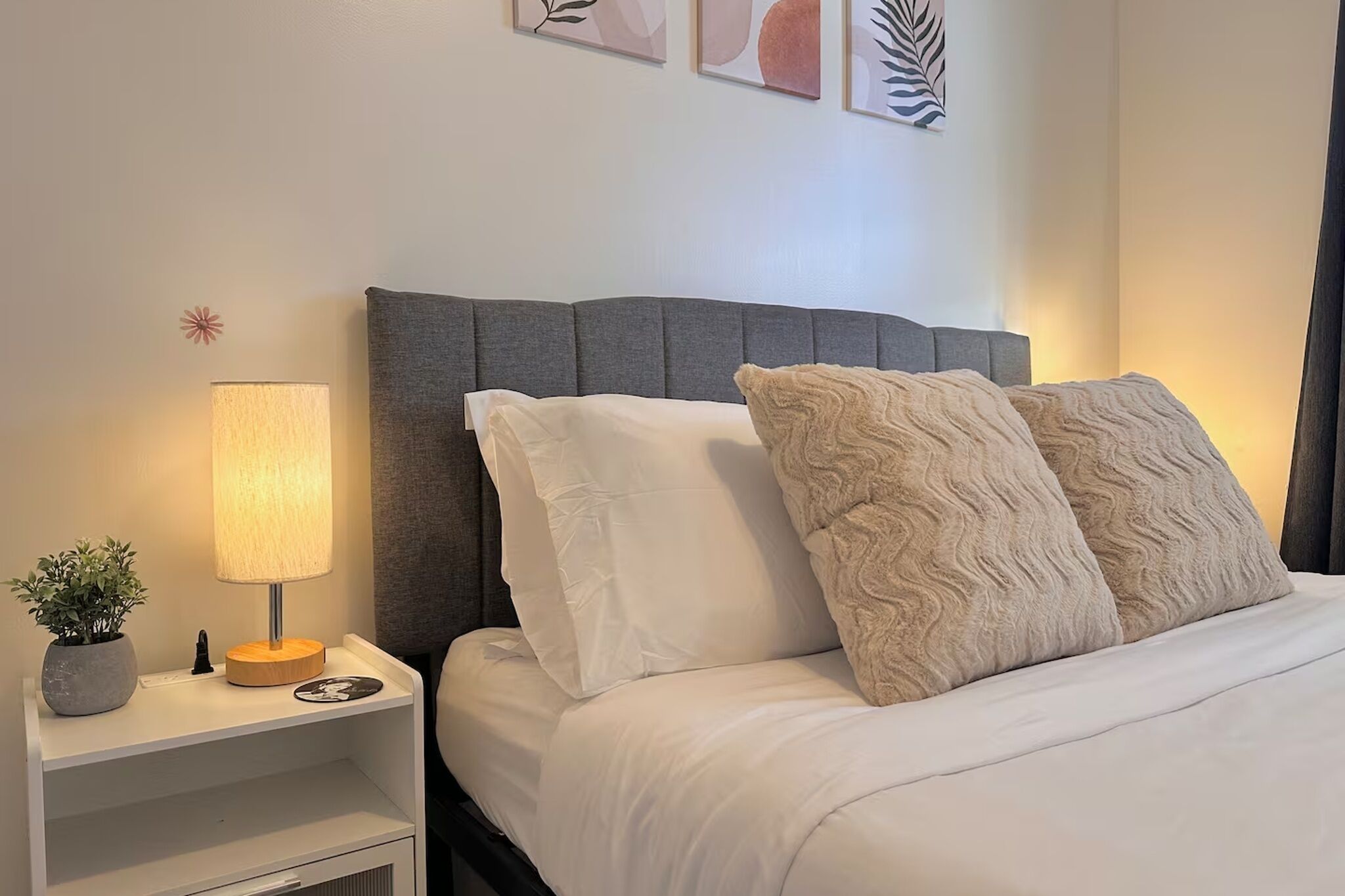 Una cama bien hecha con almohadas blancas y beige, un cabecero tapizado en gris, una mesita de noche con una lámpara, una planta pequeña y un plato decorativo.