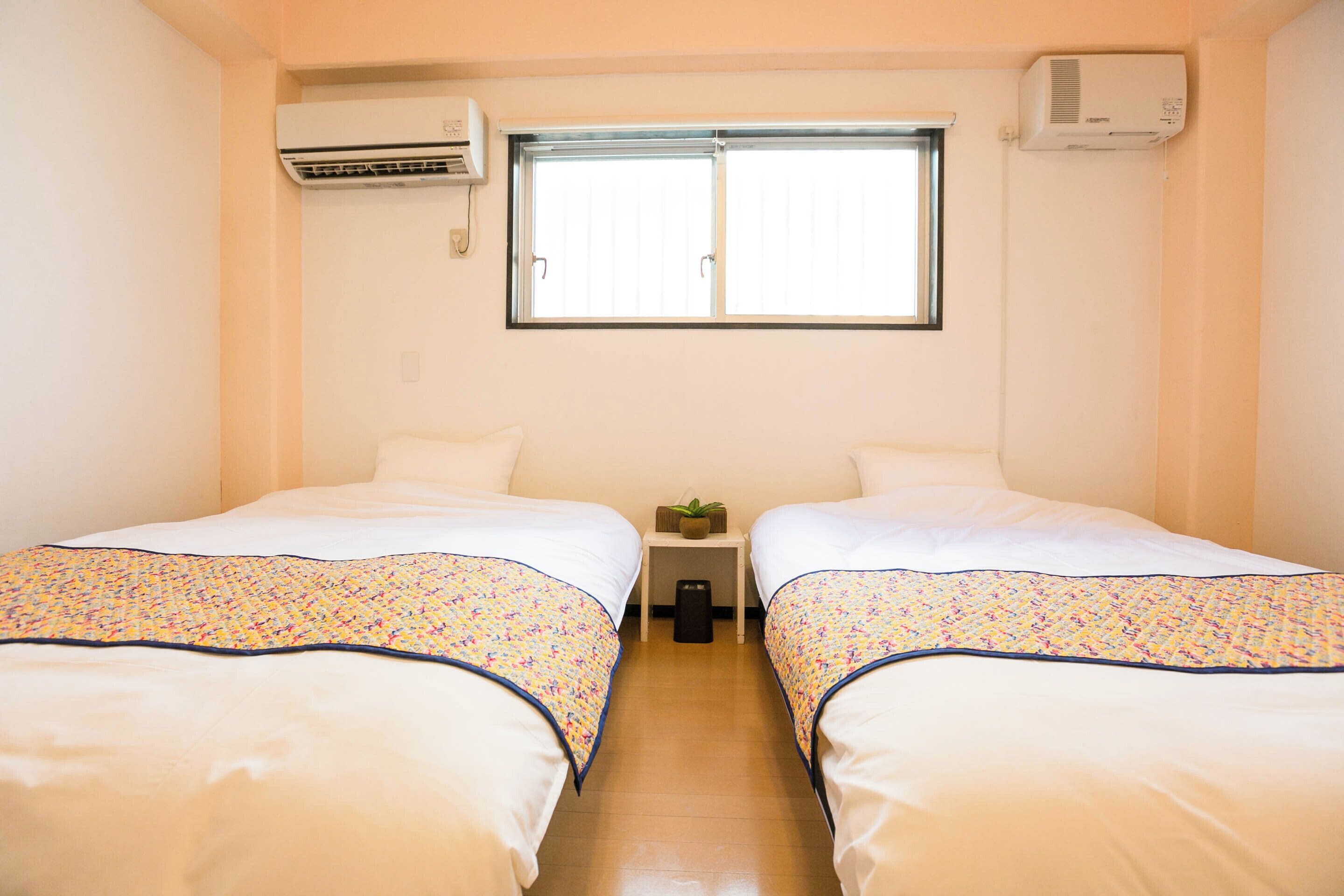 A hotel room with two beds, a window, an air conditioner, and a small table with a plant.