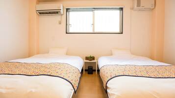 A hotel room with two beds, a window, an air conditioner, and a small table with a plant.