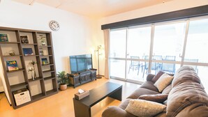 A living room with a sofa, a coffee table, a TV, a bookshelf, and a large window. - Public Inn Okinawa (Chatan)