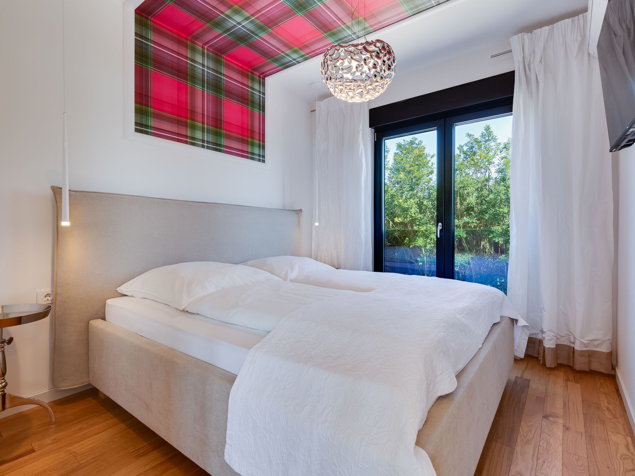 A modern bedroom with a large bed, a ceiling light fixture, and a view of greenery outside.