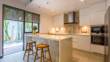 A modern kitchen with a large island, two bar stools, a stainless steel oven, and a view of a pool through large windows.