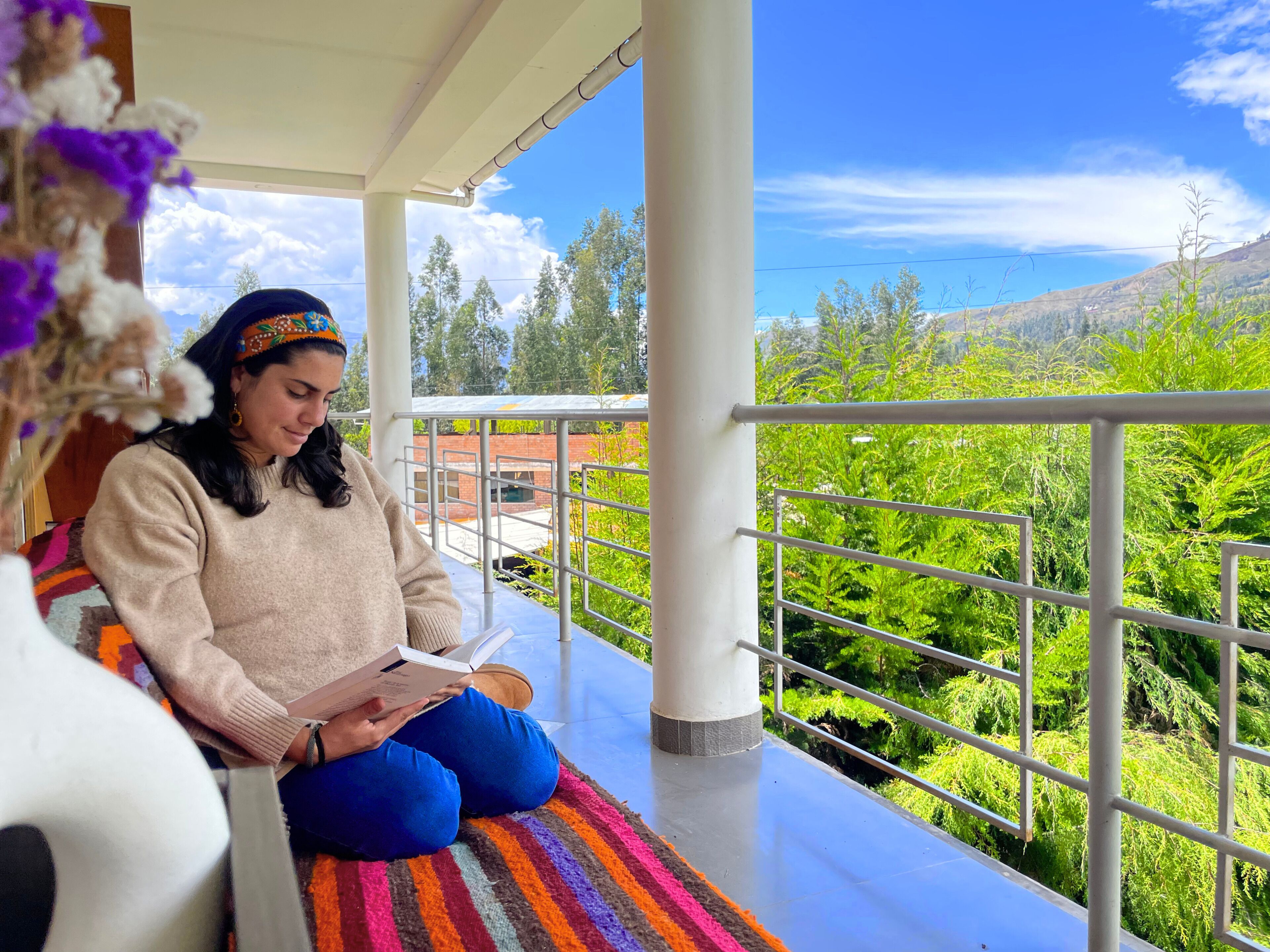 A person sitting on a colorful striped rug in a covered outdoor area with a view of green hills and a clear sky.