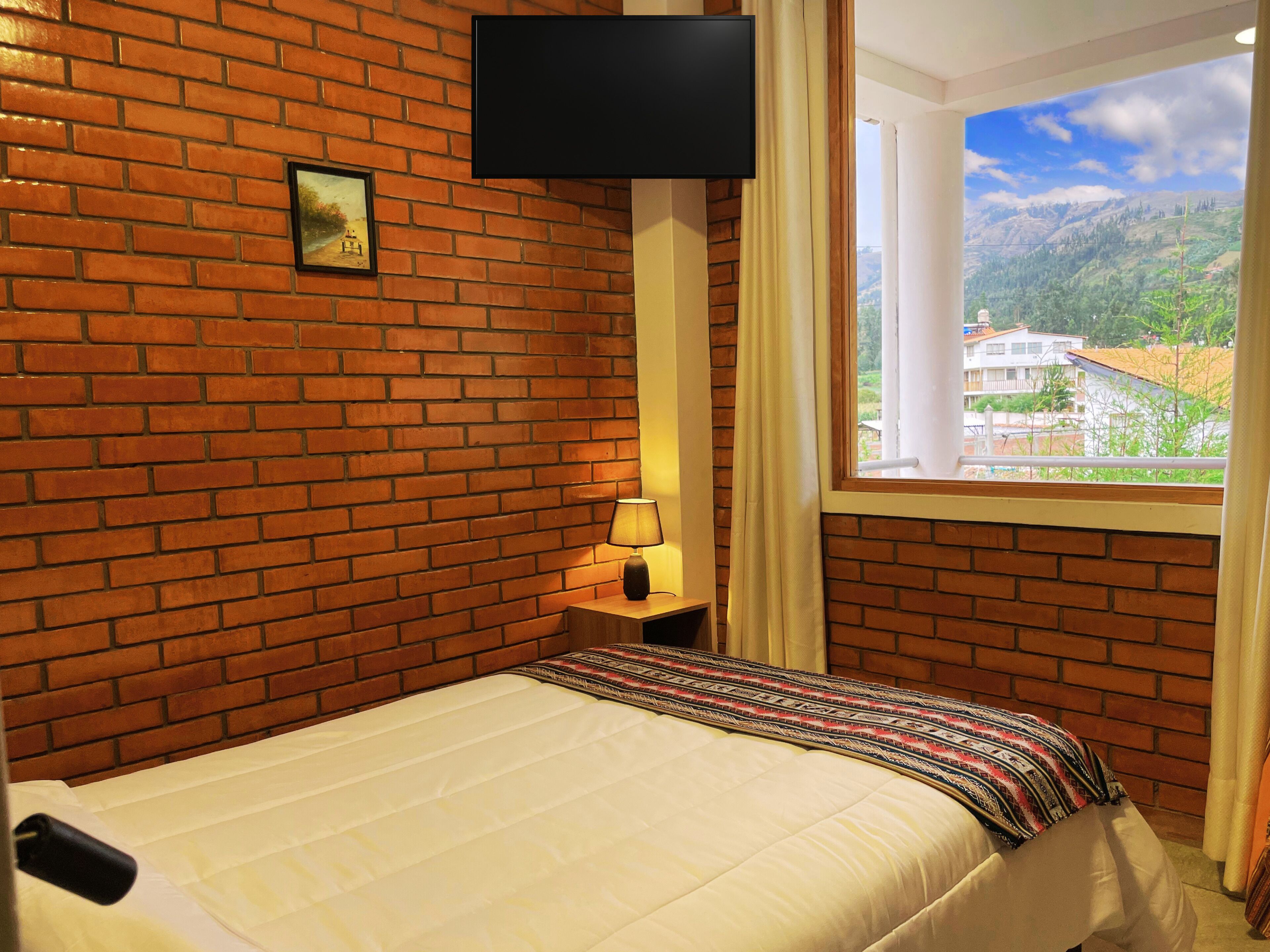 A hotel room with a brick wall, a bed with a patterned blanket, a bedside lamp, a TV mounted on the wall, and a window with a view of buildings and mountains.