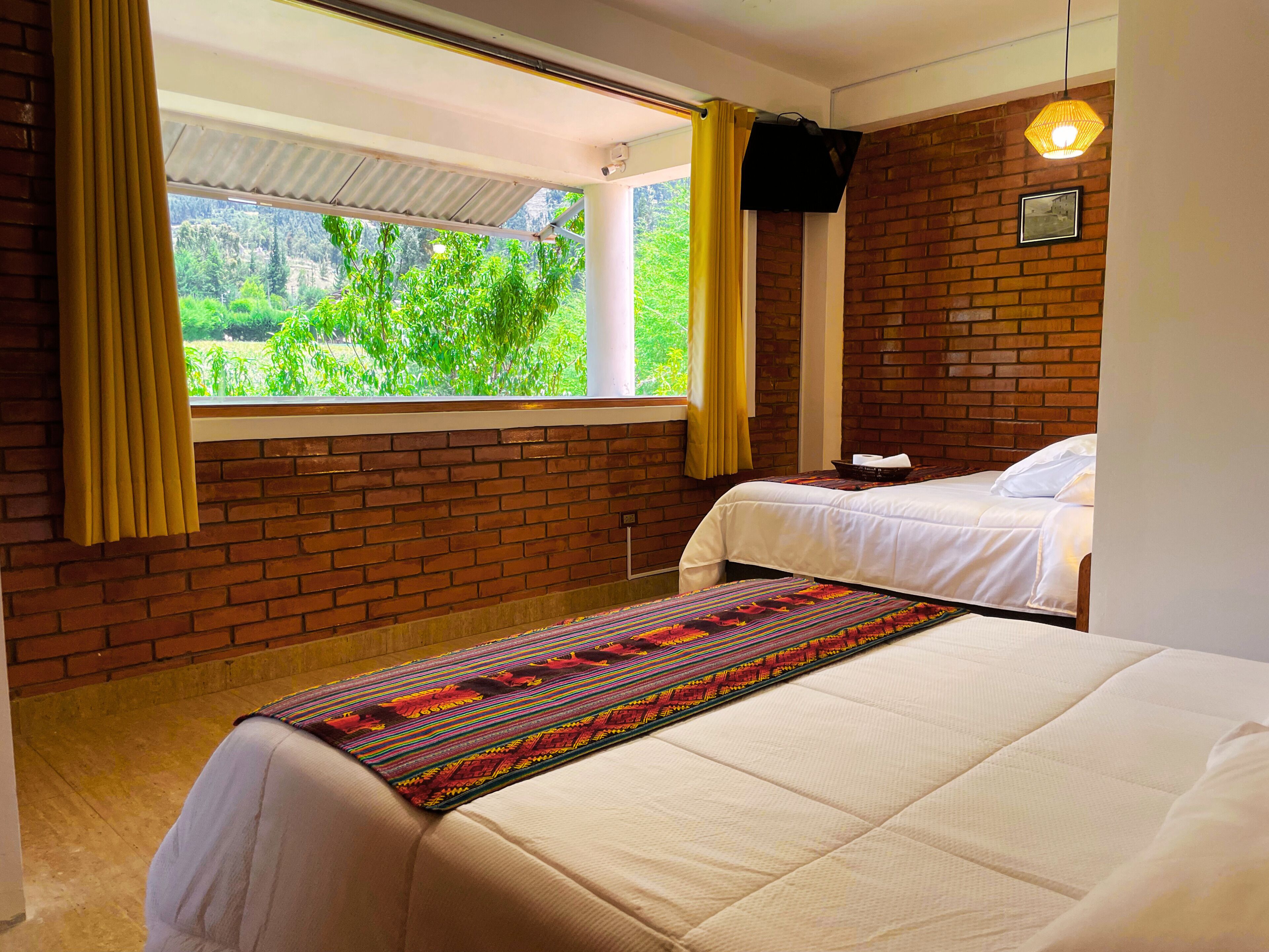 Hotelkamer met een bakstenen muur, een raam met gordijnen, een bed met een kleurrijke deken en een kleine tafel met een lamp.