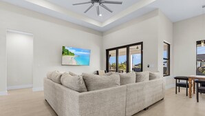 A spacious living room with a beige sofa, a dining table with chairs, and a large window offering a view of the outdoors. - Modern 4-bed home with private heated pool, outdoor bar and grill in Cape Coral! (Cape Coral)