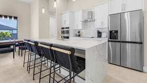 A modern kitchen with a large island, bar stools, stainless steel appliances, and pendant lighting. - Modern 4-bed home with private heated pool, outdoor bar and grill in Cape Coral! (Cape Coral)