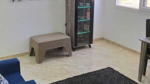 Una sala de estar con un sofá azul, una alfombra negra, un televisor montado en la pared y un aparador con puertas de vidrio.