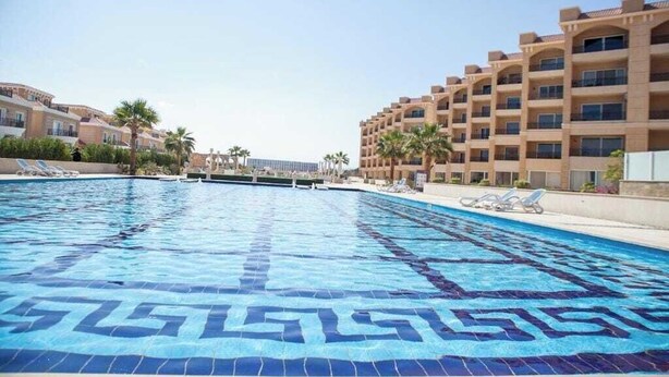 Una piscina con un diseño geométrico de azulejos, rodeada de sillones de descanso y palmeras, con un edificio de varios pisos al fondo.