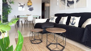 Un salon moderne avec un canapé noir, deux tables hexagonales, un coin repas avec une table et des chaises, et des œuvres d’art murales représentant des baleines et des ancres.