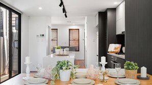 A modern dining area with a wooden table set for four, white chairs, and a kitchen in the background. - Newtown Family Stay | 6 Beds + Ducted Aircon (Newtown)