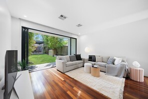 A modern living room with a large sliding glass door leading to a backyard, a grey sofa set, a coffee table, and a TV. - Newtown Family Stay | 6 Beds + Ducted Aircon (Newtown)