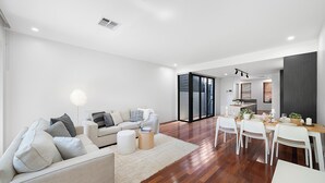 A modern living room with a sofa, coffee table, and a dining area with chairs and a table. - Newtown Family Stay | 6 Beds + Ducted Aircon (Newtown)