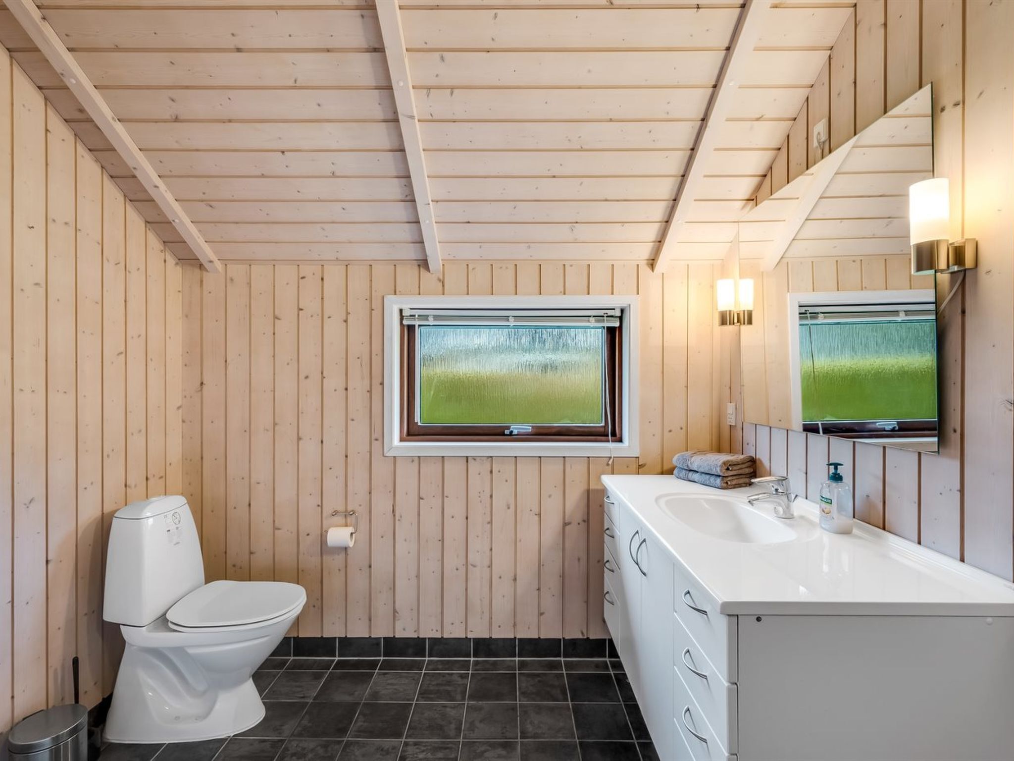 A modern bathroom with wooden walls, a white toilet, a large window with a view, a white sink, and a mirror.