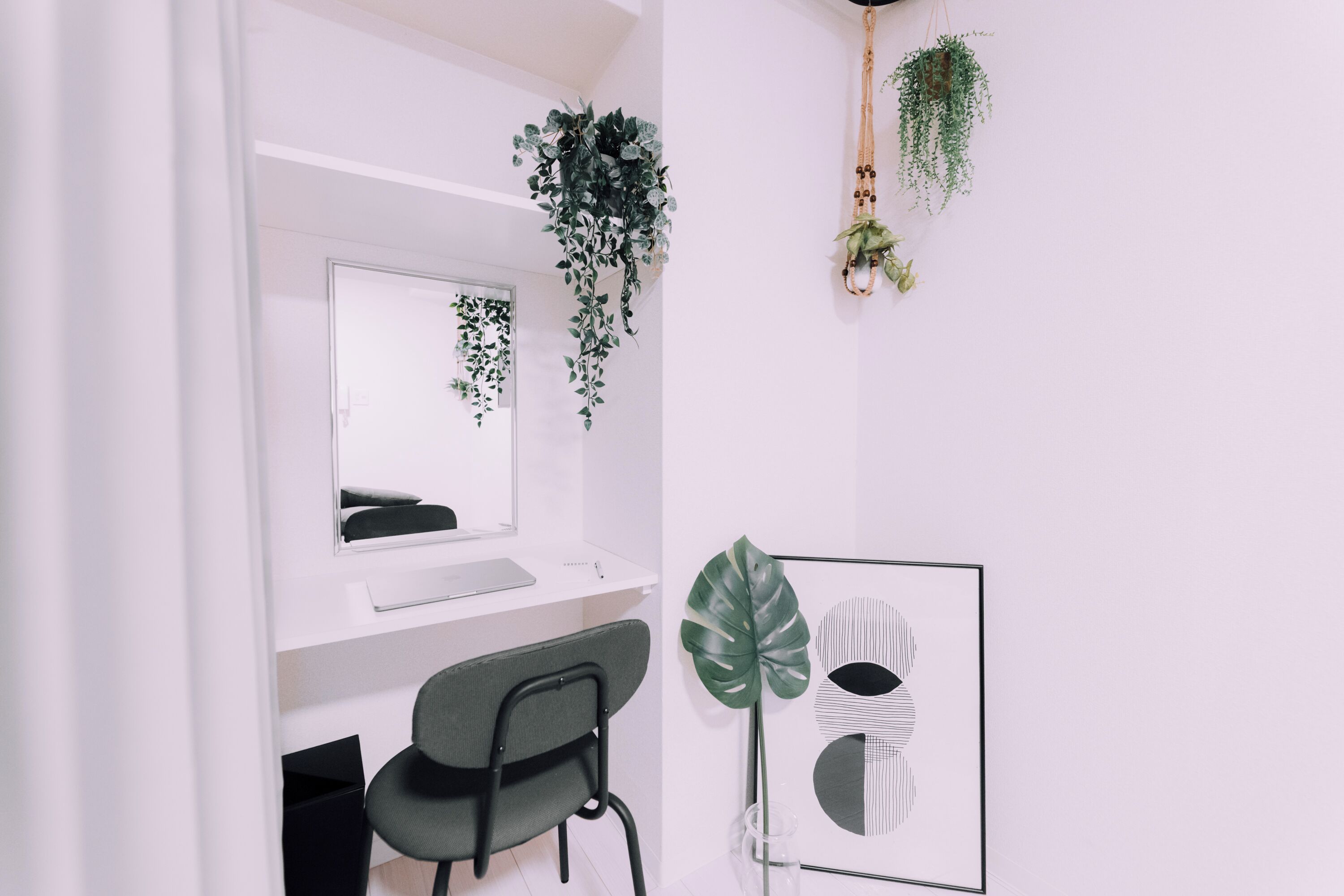 Ein modernes, minimalistisches Zimmer mit weißem Schreibtisch, einem Stuhl, einem großen Spiegel und hängenden Pflanzen.