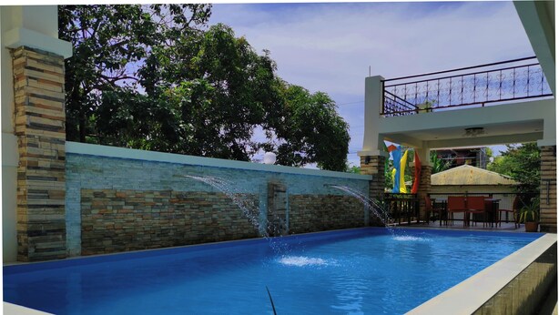 Una piscina con muro in pietra e elemento decorativo acquatico. È presente una zona relax coperta con ombrelloni e sedie.