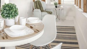 Une pièce chaleureuse avec un plafond en bois, une table à manger dressée avec de l’assiette blanche et des couverts en argent, un canapé blanc et une petite table sur laquelle est posée une plante.
