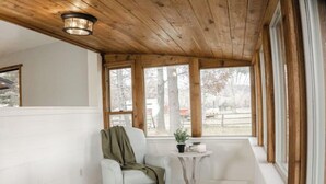 Une pièce chaleureuse avec un plafond en bois, un fauteuil blanc agrémenté d’un plaid vert, une petite table avec une plante et un tapis à rayures.