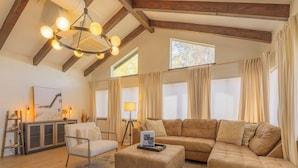 Un soggiorno spazioso con un ampio divano modulare, una poltrona e un tavolino. Pavimento in legno, lampadario e una grande finestra con tende.