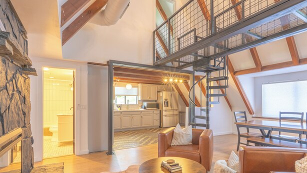 Un loft spazioso con soffitto alto, travi in legno a vista, una parete d’accento in pietra, un’area salotto accogliente con divani in pelle, un tavolo da pranzo e una cucina sullo sfondo.