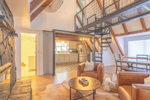 Un loft spacieux avec de hauts plafonds, des poutres apparentes, un mur d’accent en pierre, un coin salon confortable avec des canapés en cuir, une table à manger et une cuisine à l’arrière-plan.