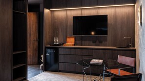 Una cocina moderna con armarios de madera oscura, un televisor empotrado, un refrigerador negro y un pequeño comedor con mesa de vidrio y sillas de metal.
